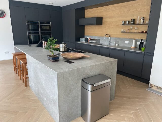 Contemporary open-plan kitchen with waterfall stone island, dark cabinetry and oak wall panelling by Tolle Kitchens